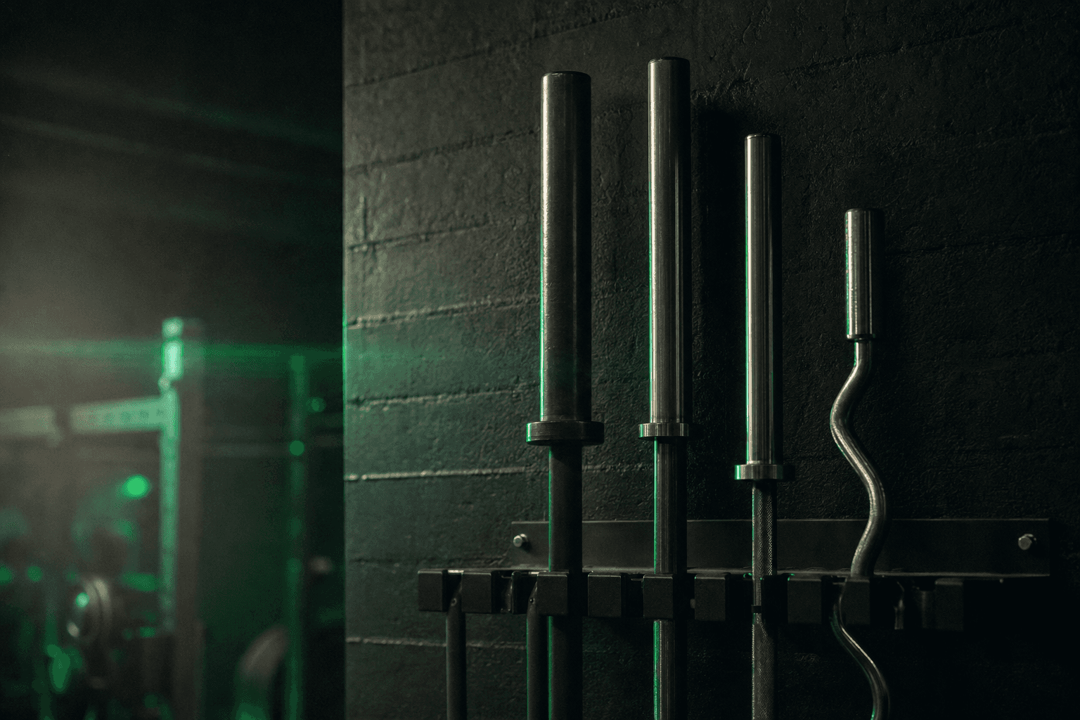 Row of different barbells racked against a dark gym wall, from thick powerlifting bar to curved EZ curl bar