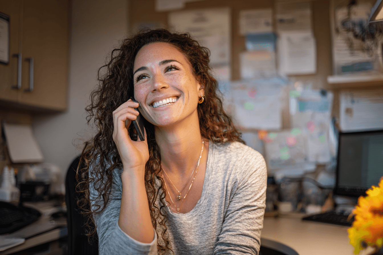 a woman in her 30s, on her cell phone and smiling at her desk in a home care agency office.