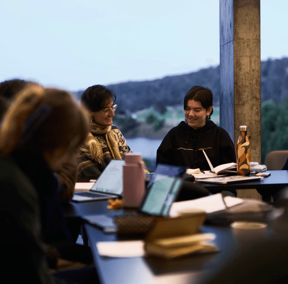 A group of young adults sit around a long table in a bright room with large windows, engaged in discussion. Laptops, notebooks and water bottles are scattered across the table, and several participants smile and lean toward one another, with a forested landscape visible outside.