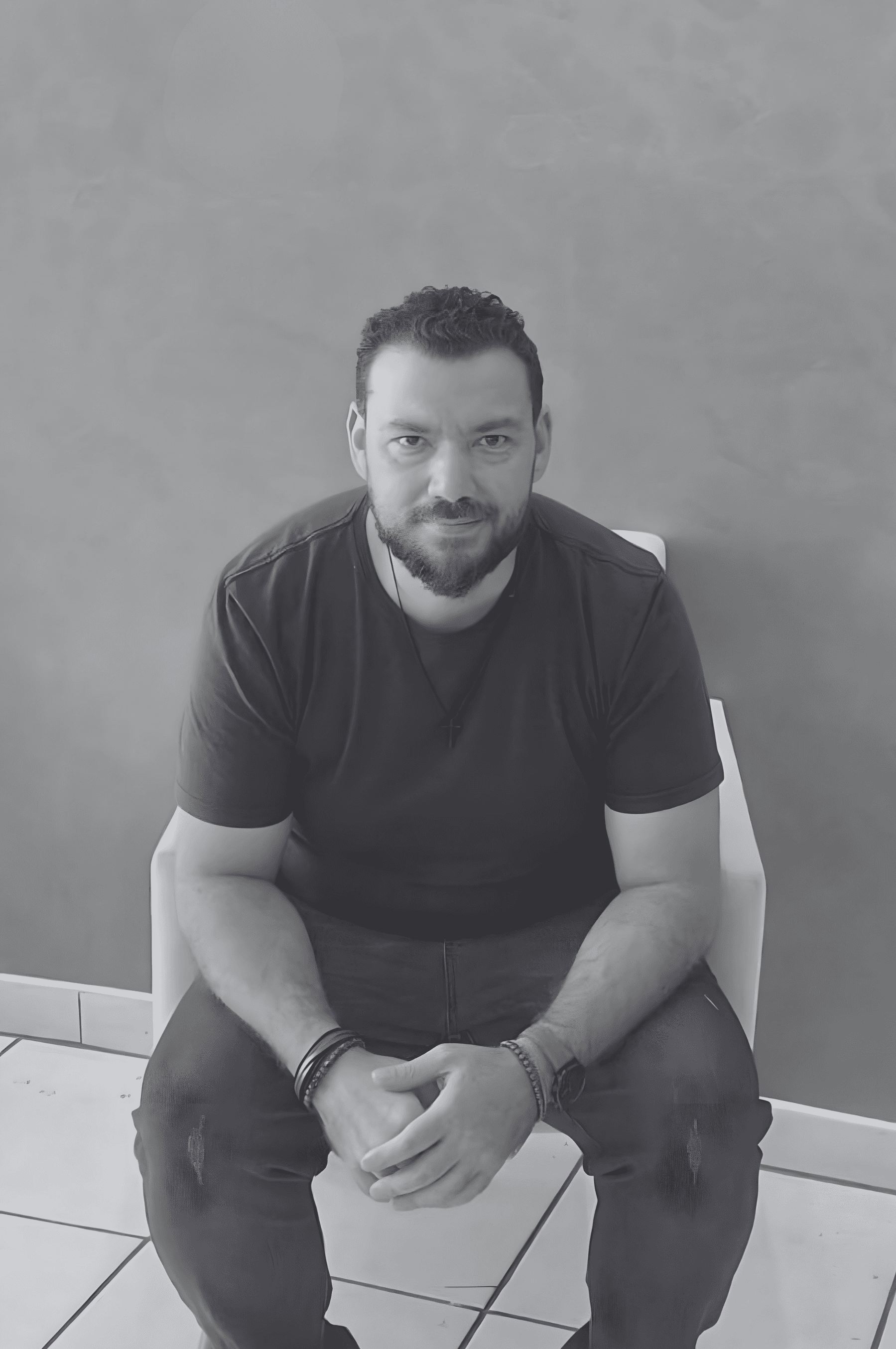 A person with curly hair and glasses smiles at the camera, seated in a cozy workspace with warm lighting.