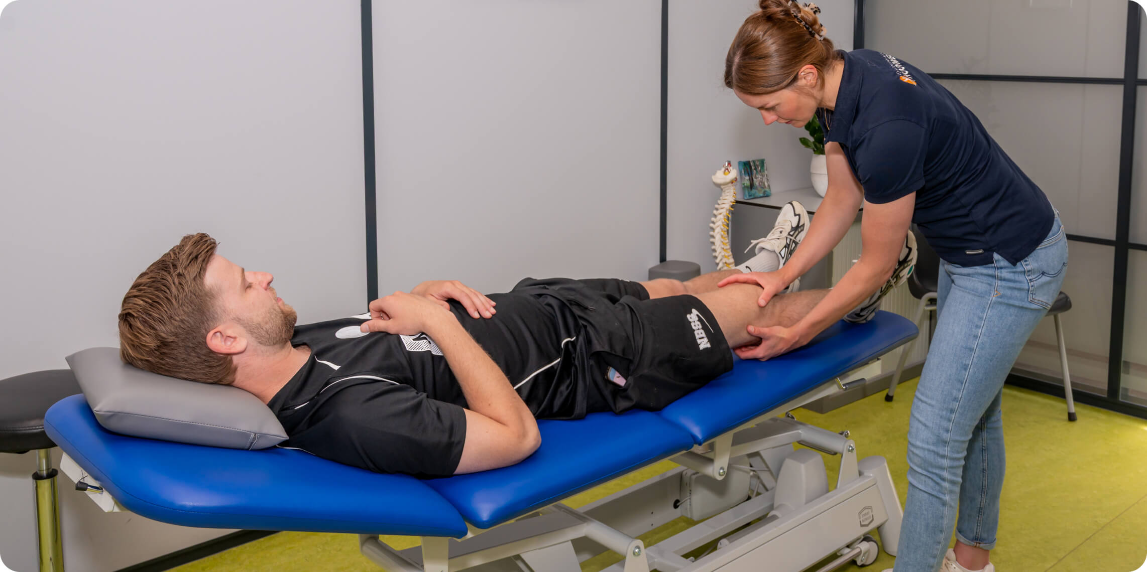 A physiotherapist in a navy shirt is examining the leg of a male patient lying on a blue padded treatment table in a modern clinic room, with a spine model visible in the background.