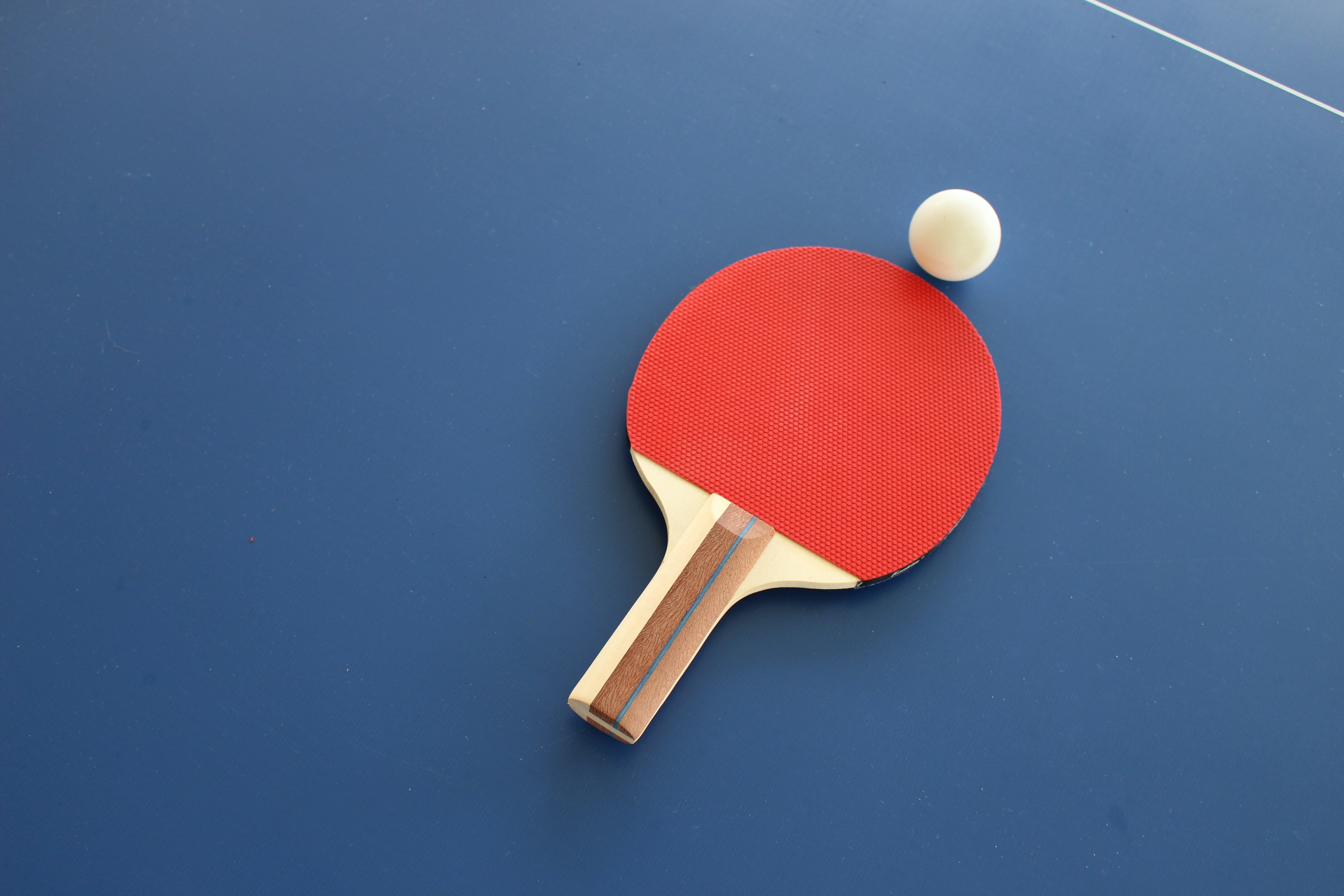 A ping pong paddle with a red rubber surface and a wooden handle is resting on a blue ping pong table, with a white ping pong ball hovering above it.