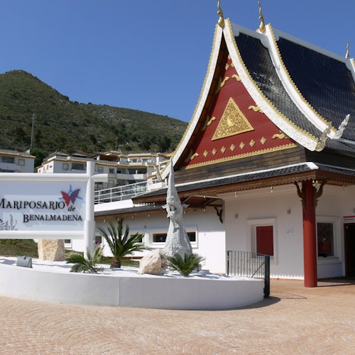 Un edificio de estilo tailandés con un letrero que dice "Mariposario de Benalmádena" y una estatua al frente, situado contra una ladera.
