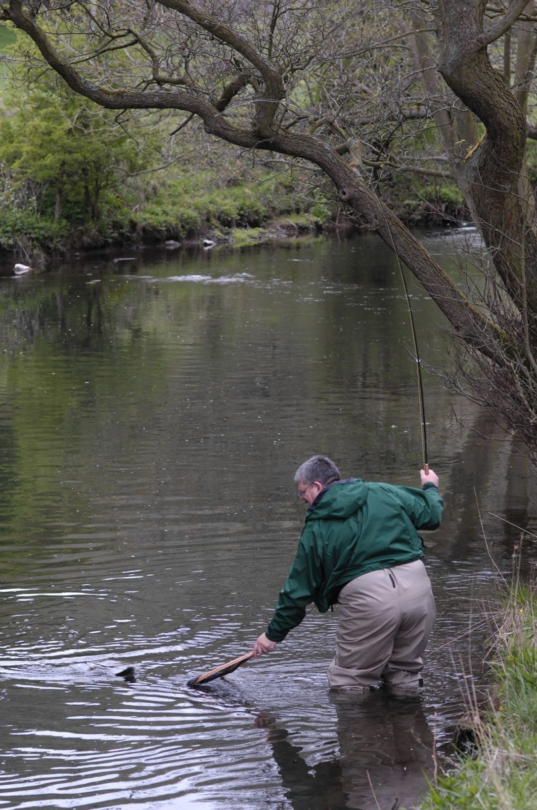 A man stands by a river, casting a fishing line, surrounded by trees and the gentle flow of water.