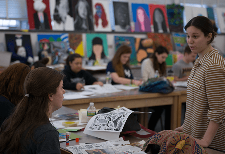 A woman with dark brown hair tied in a ponytail sits facing left, wearing a dark shirt. To her right, a woman with dark hair in a striped shirt stands facing forward, looking toward the seated woman. Behind them, several other people are seated at a table covered with papers and art supplies, including water bottles and a blue bag. Behind the table, a wall displays a row of colorful artworks featuring black and white portraits.