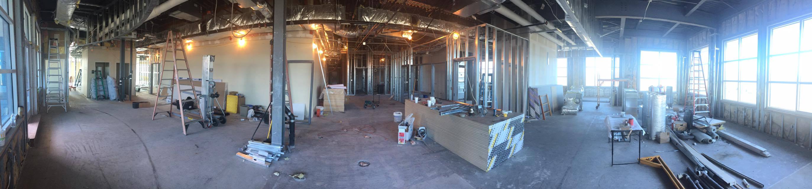 A spacious room with exposed beams and columns and large windows, filled with natural light and under construction.