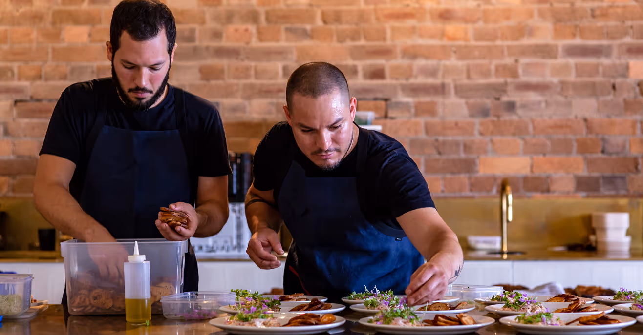Two chefs in aprons plating canapes along a kitchen bench in front of a brick wall