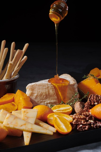 Charcuterie board with thin breadsticks, wedges of clementines, orange-and-white marbled sliced cheddar, halved kumquats, pecan halves, mimolette cheese, dried apricot halves, and a large wedge of brie being drizzled from above with honey from a wooden honey dipper.