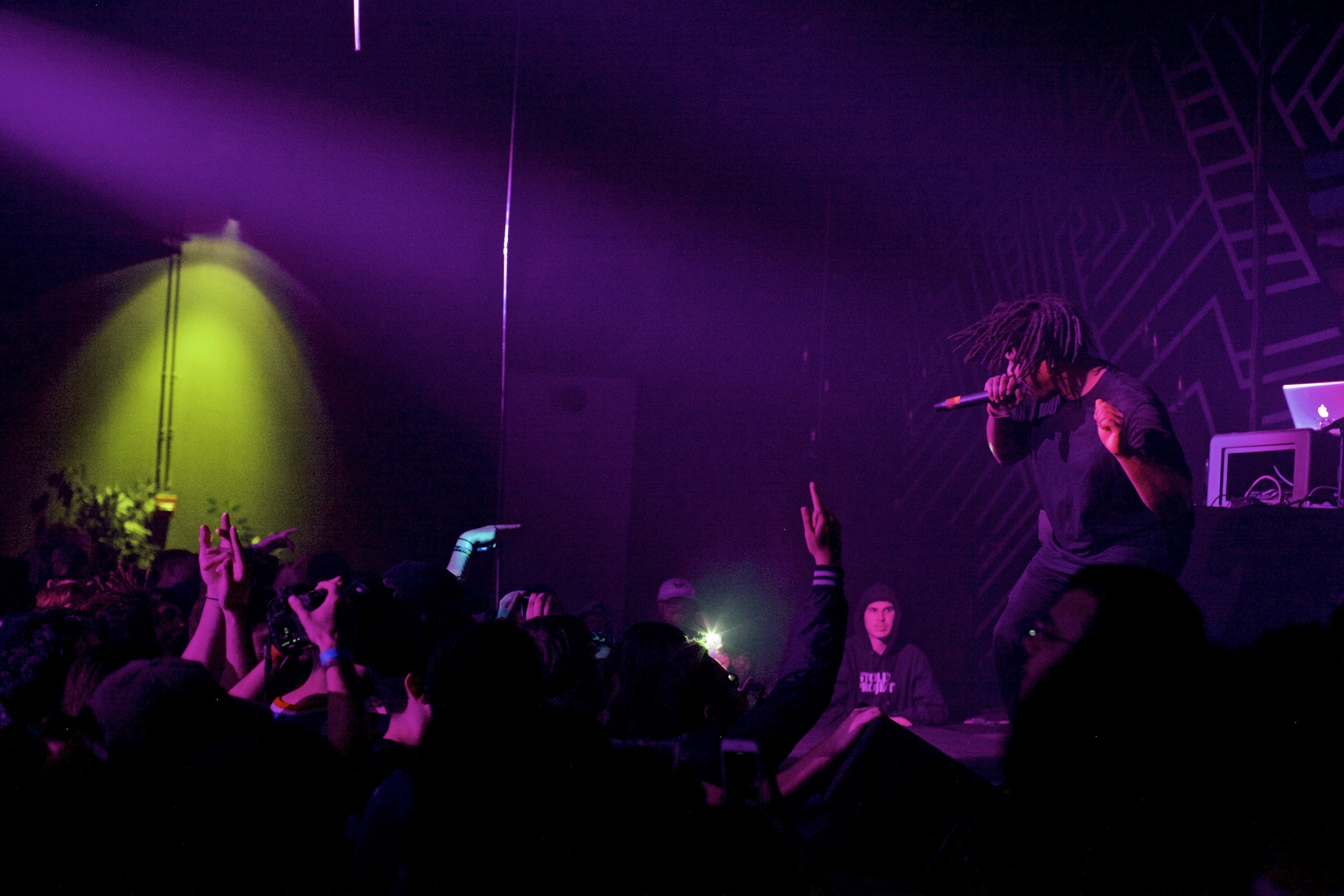 Rapper performing onstage in purple lighting with audience hands raised in the foreground.