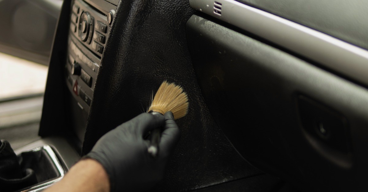 A close-up of a person wearing black gloves holding a soft tan brush to clean the dashboard of a car.