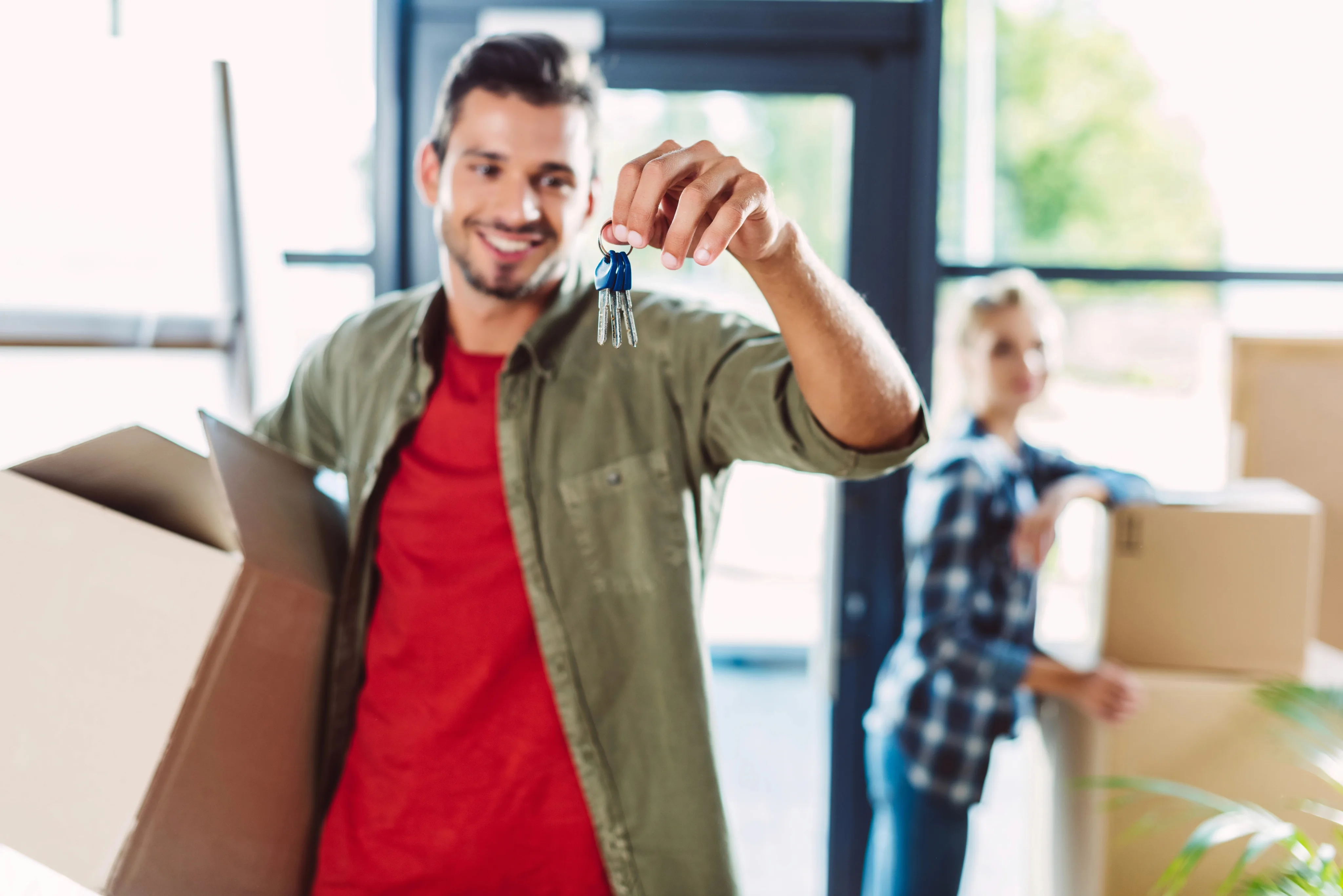 A smiling man in a red shirt and olive jacket proudly displays house keys with a blue keychain while standing in a bright, modern home with moving boxes and another person in the background.