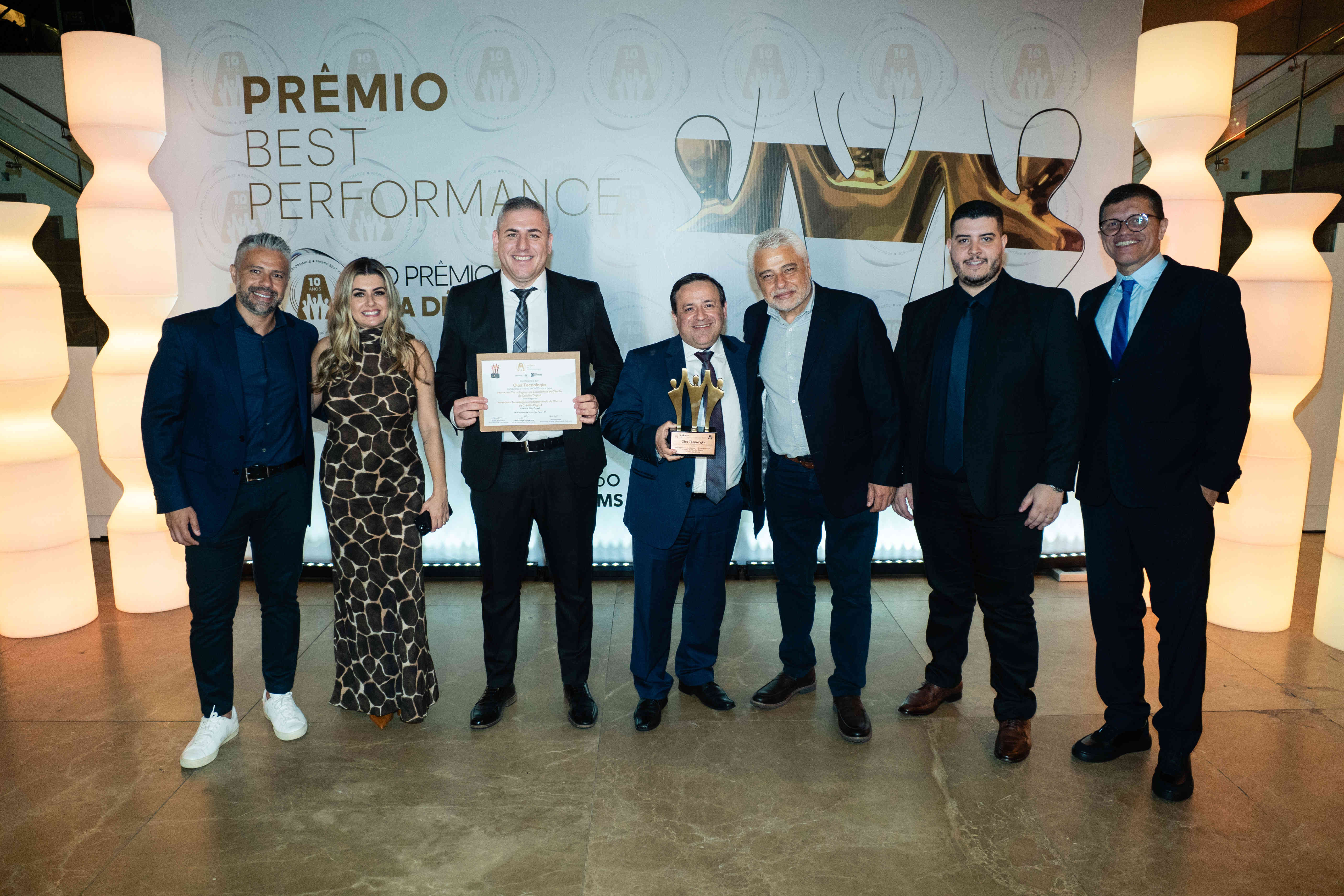 Executivos da Olos Tecnologia posando com troféu bronze e certificado no palco do Prêmio Best Performance 2025