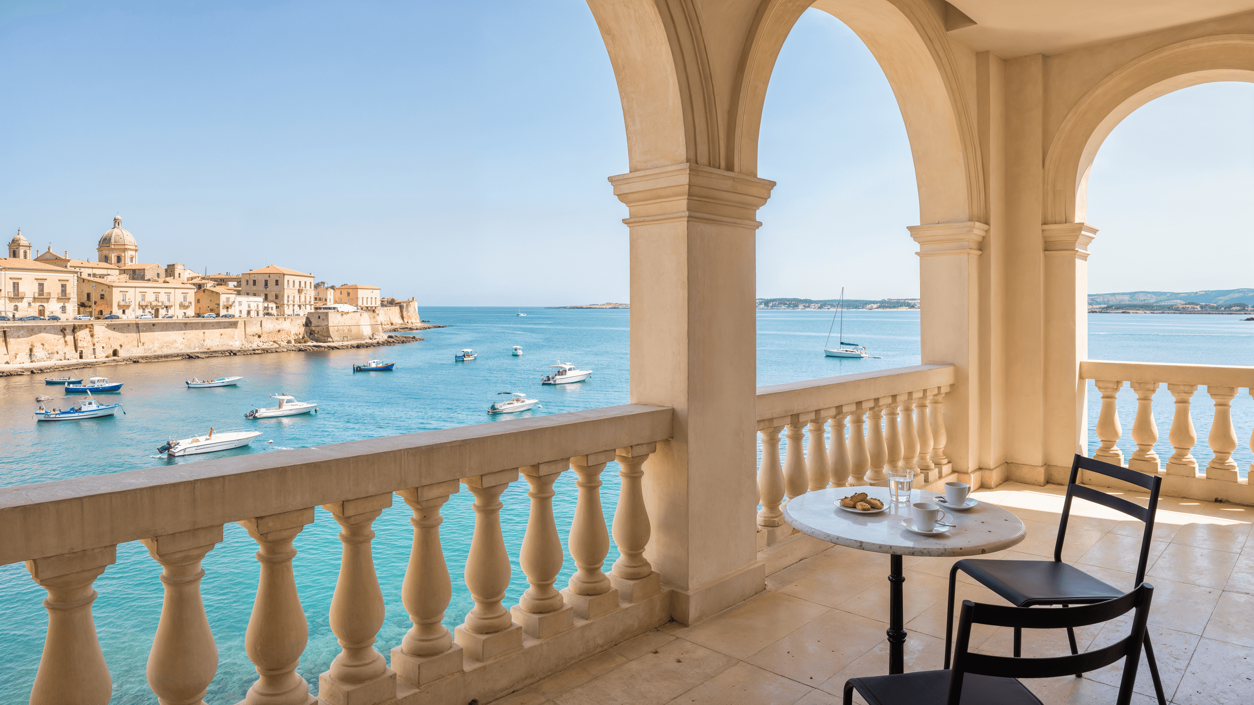 Sunlit arched loggia with a stone balustrade and café table overlooking a Mediterranean harbor town