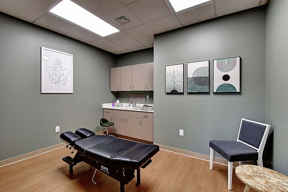 Modern therapy room with gray walls, a massage table, a chair, and minimalistic decor. Bright and inviting atmosphere.