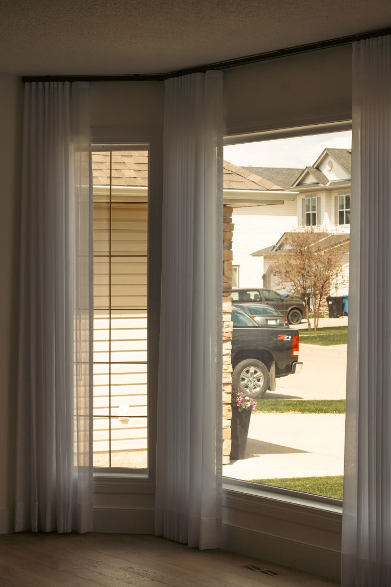 Image of Sheer curtains frame a large window with a view of a suburban street and parked truck outside.