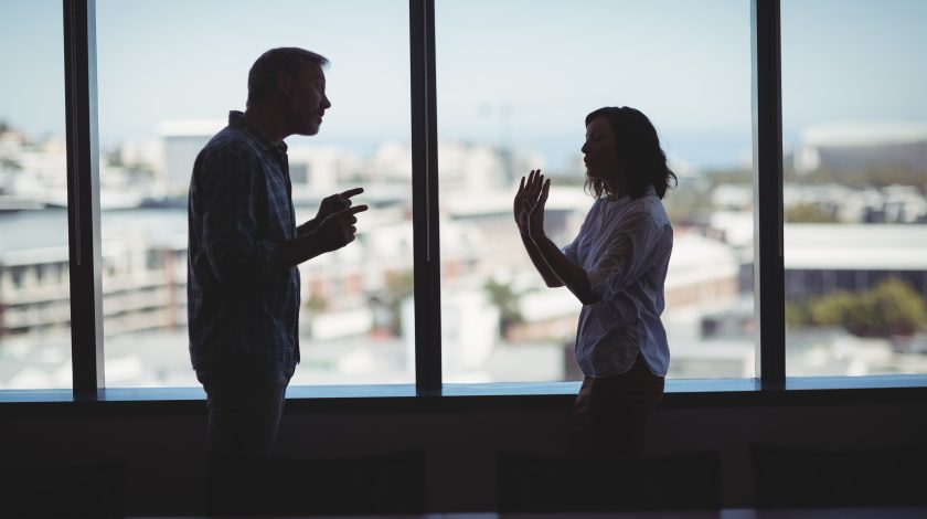 A silhouette of a man and woman engaged in a heated discussion is set against a large window overlooking a blurred cityscape, conveying tension and urban lifestyle contrast.