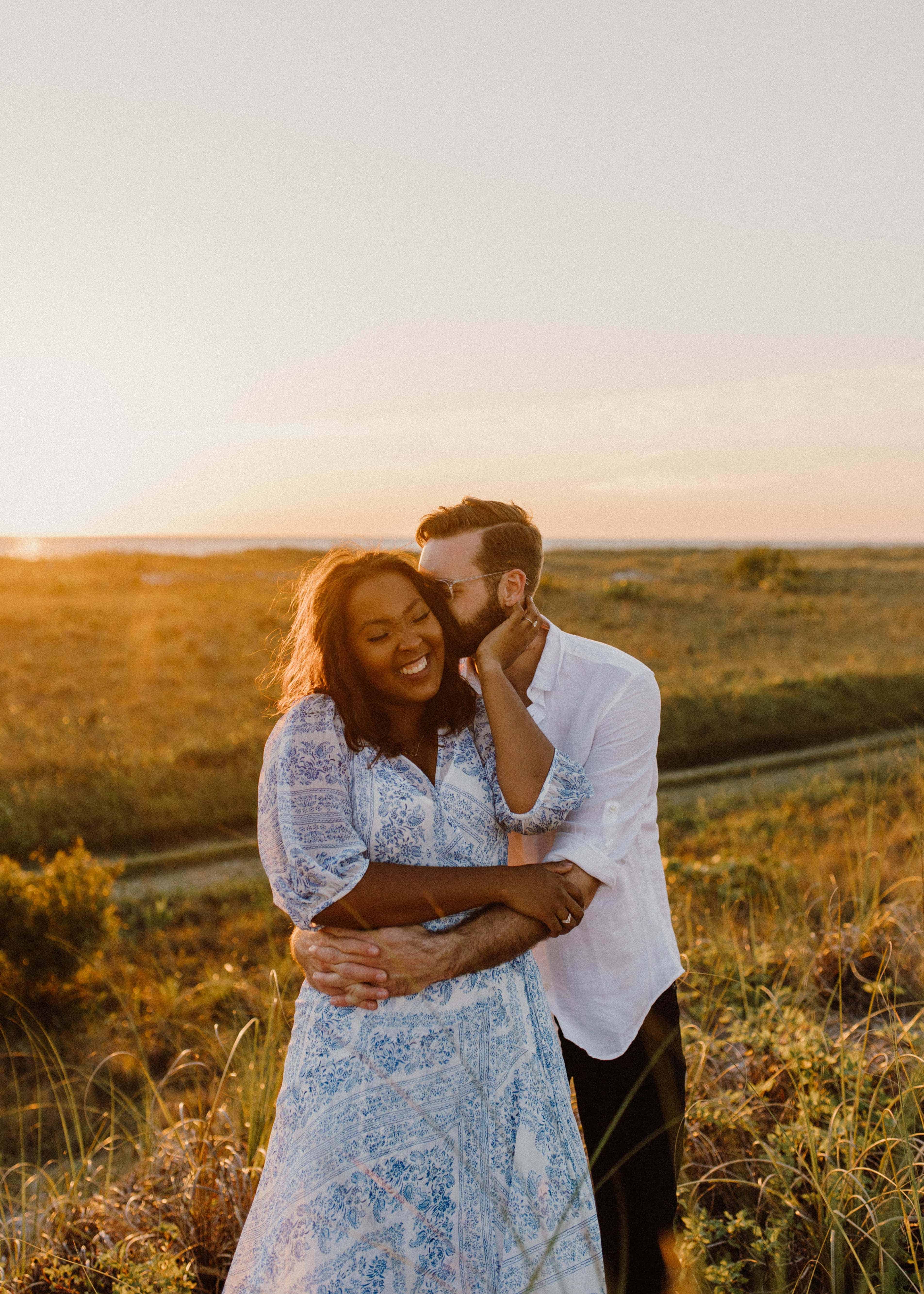  A photo of a couple hugging in a grassy field at sunset. The man is wearing a white shirt and the woman is in a blue and white dress.