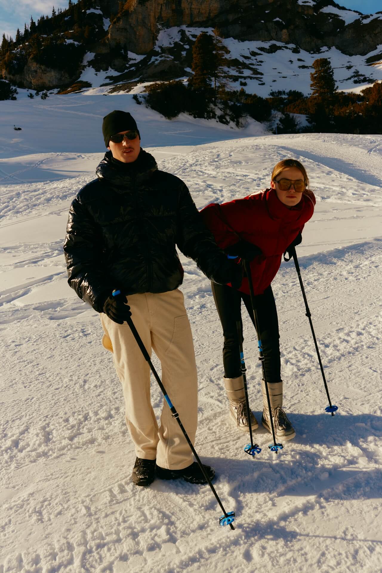 Two people in winter clothing stand on a snowy slope with ski poles, surrounded by trees and rocky hills in the background, exuding a playful mood.