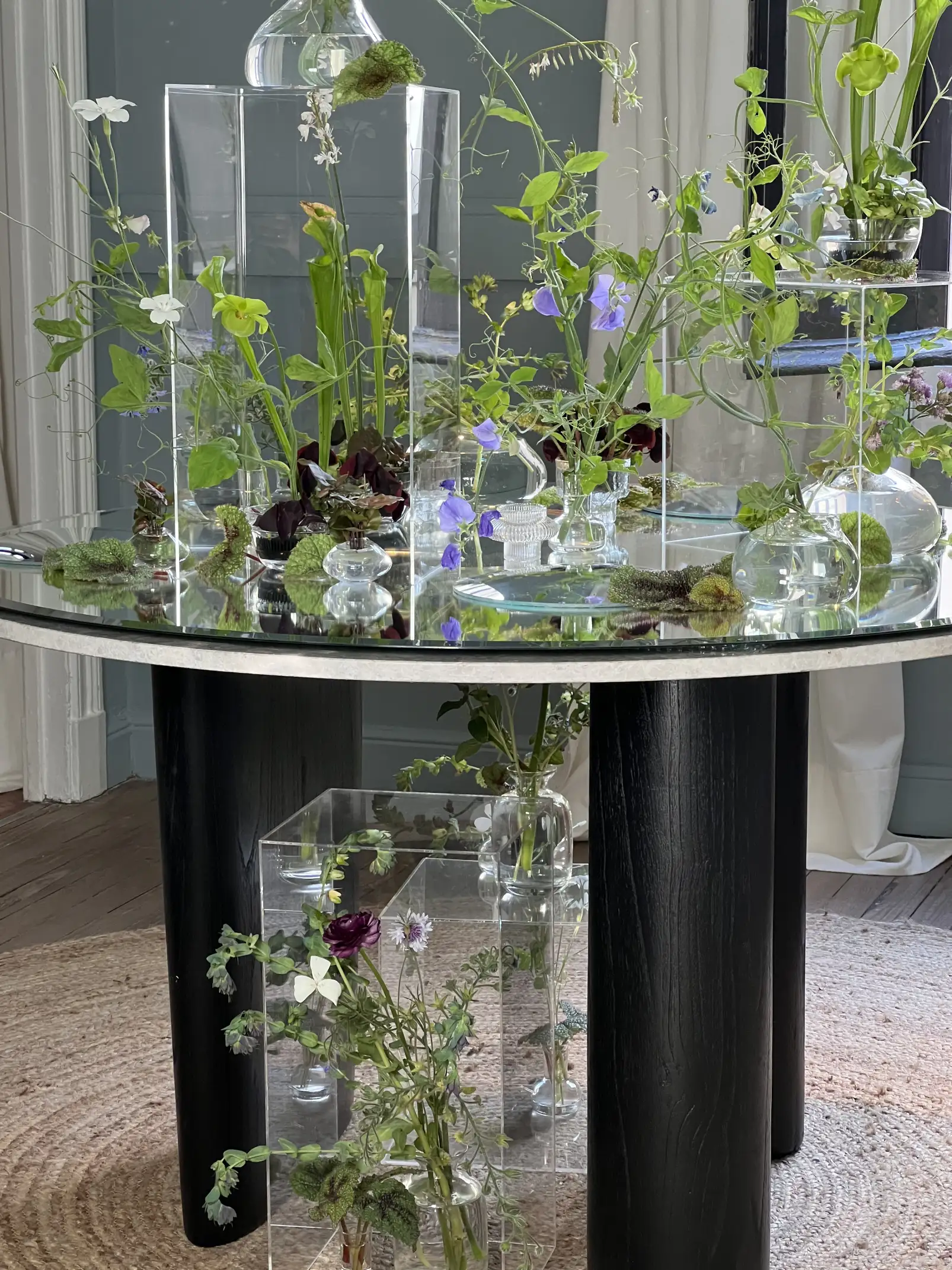 Mesa circular con bases acrílicas transparentes y múltiples arreglos florales delicados en tonos verdes y lilas.