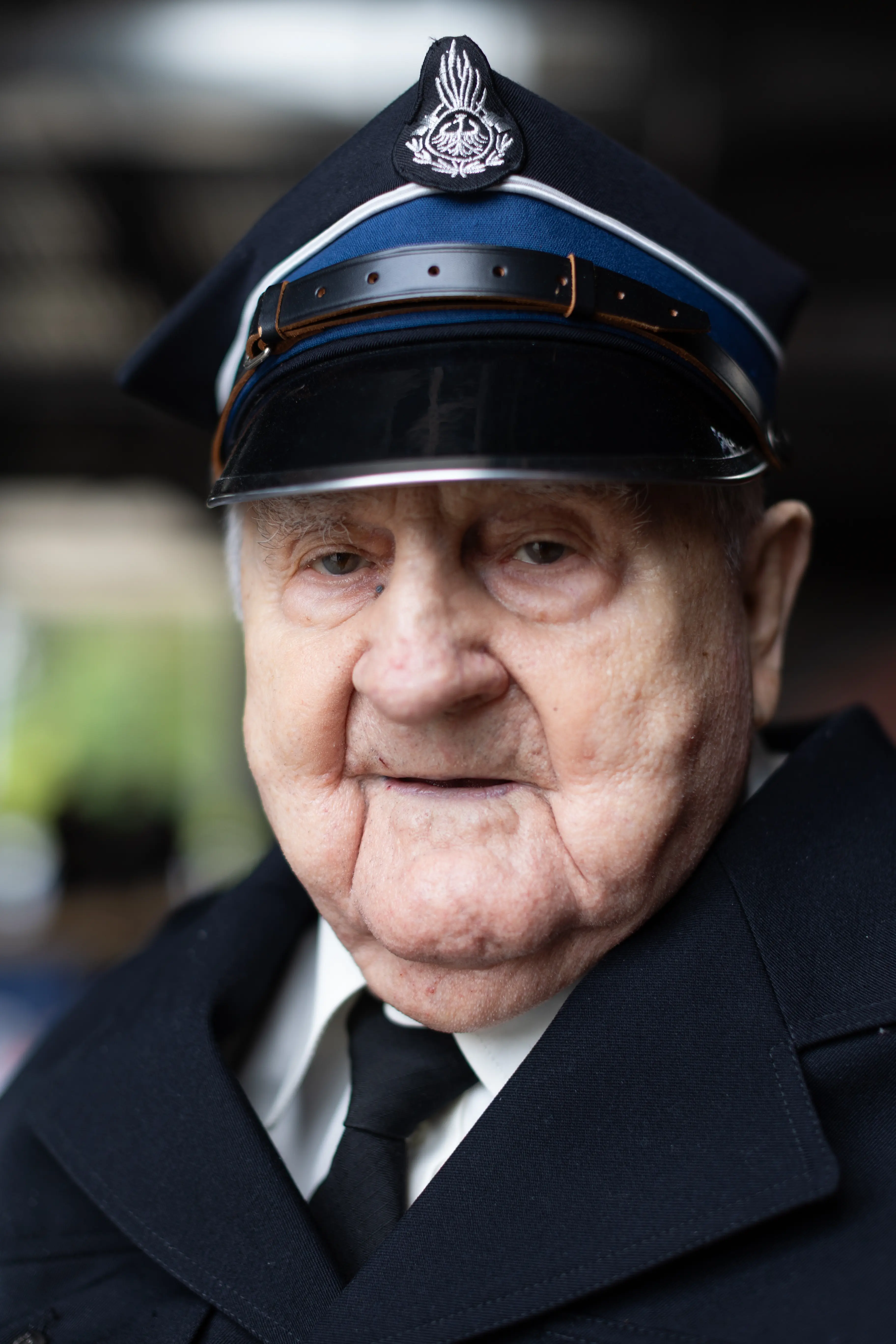 Starszy mężczyzna w czapce z munduru funkcjonariusza, patrzący prosto w obiektyw. Ubrany jest w garnitur i czarny krawat, tło jest rozmyte.