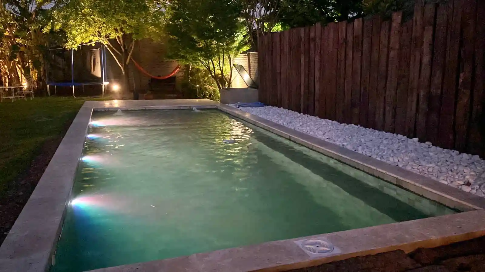 Pileta de jardín iluminada de noche con modernas luces LED subacuáticas, enmarcada por un cerco de madera rústico y un sendero de piedras decorativas blancas.