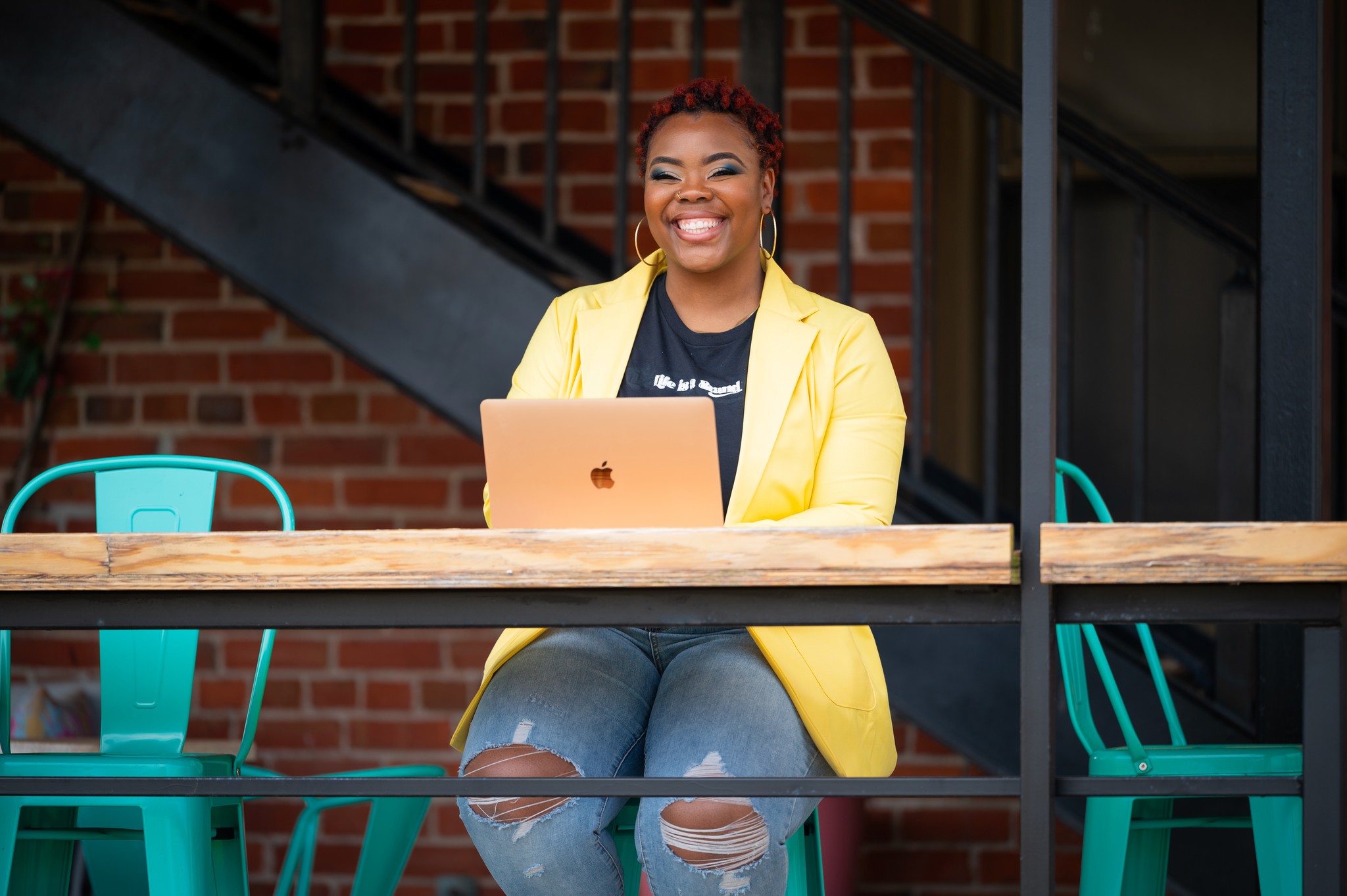 A young person with curly hair smiles at the camera while sitting in front of a laptop against a dark background.