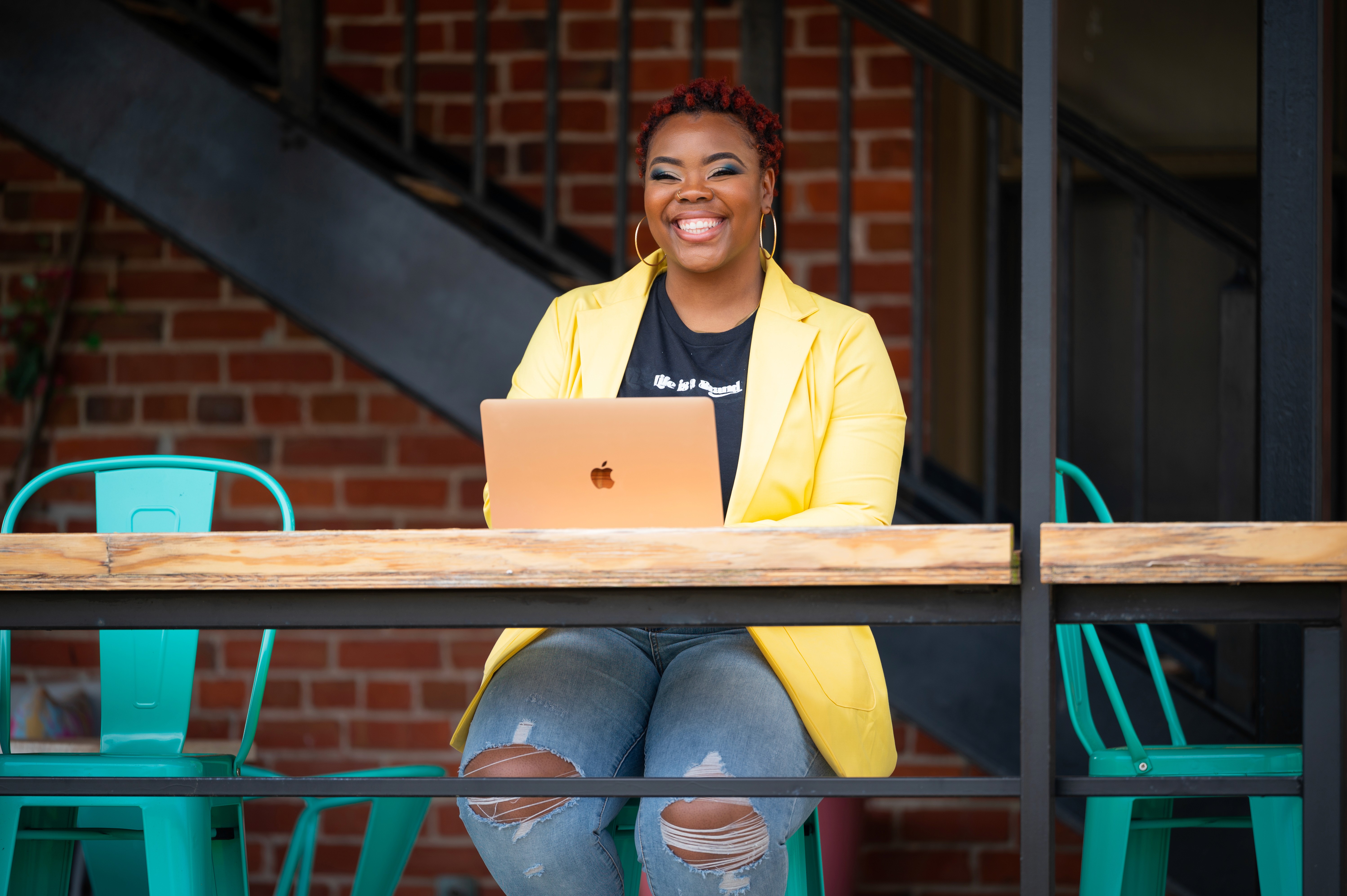 Tre'elle Tolbert sitting at her Macbook smiling outdoors wearing a life is a brand t-shirt and yellow blazer
