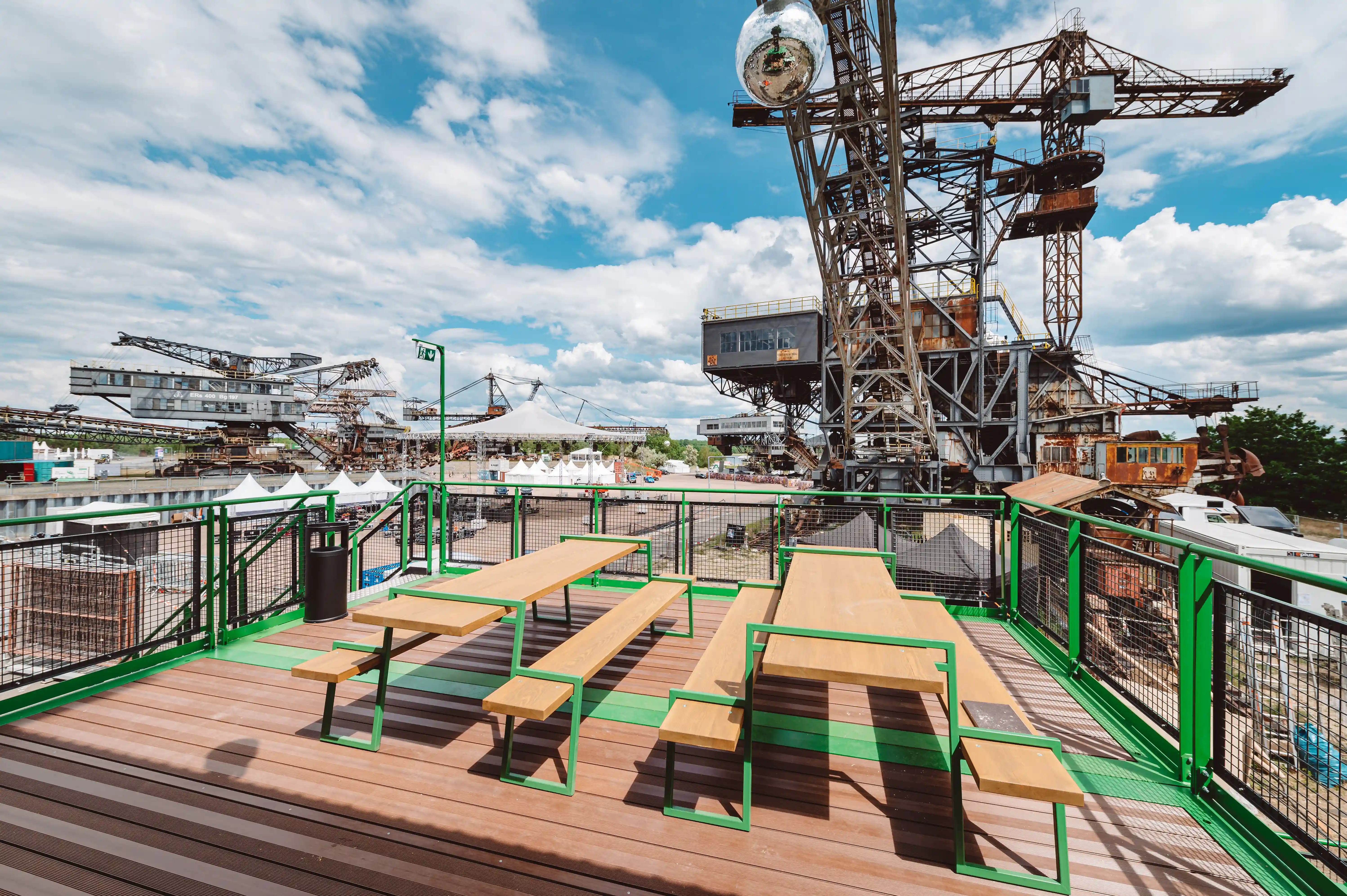 Roof terrace of the KROMBACHER stand with wooden beer table sets behind green mesh railings and a disco ball, overlooking the Ferropolis machinery.
