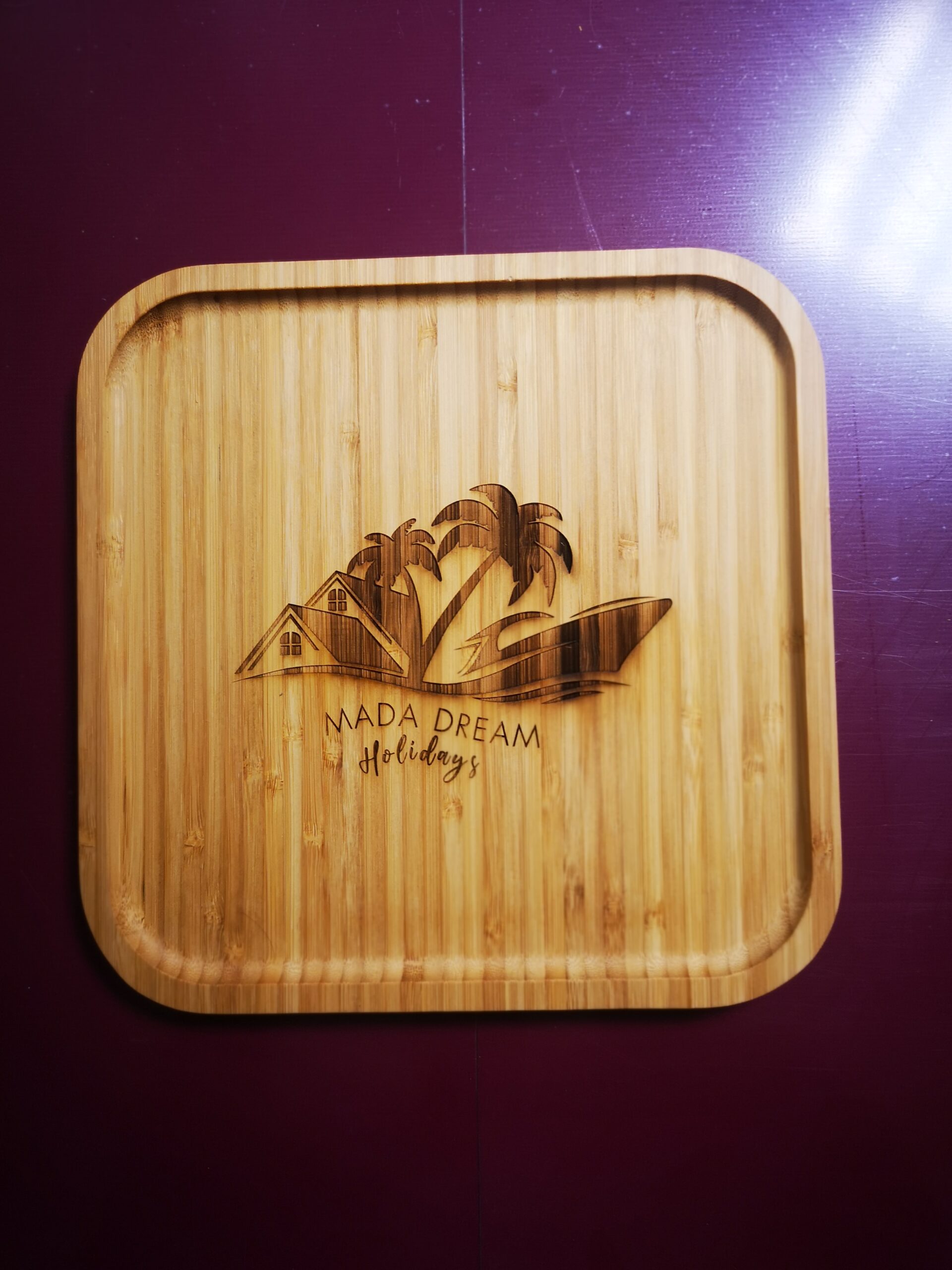 Planche à découper en bambou gravée de palmiers, d'une maison et des mots « Mada Dream Holidays », sur un fond violet intense, évoquant une ambiance tropicale.