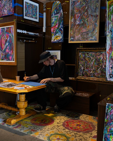 Zdjęcie przedstawiające arystę, który maluje obraz na swoim stanowisku targowym, wśród obrazów.