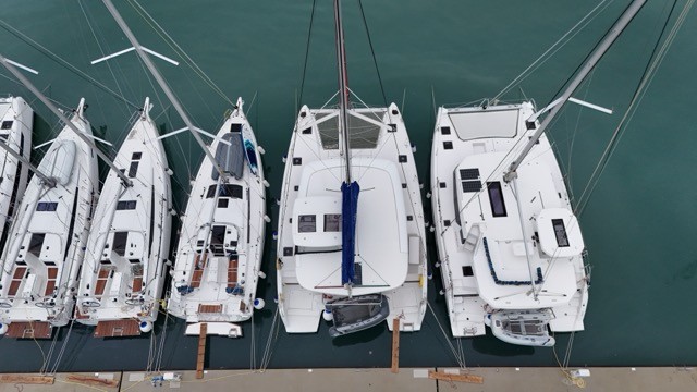 Direct overhead aerial view of a Lagoon 42 sailing catamaran berthed stern-to at a marina pontoon, showing the full deck layout including the wide bridgedeck, twin helm stations, trampolines, mast with blue halyard, and surrounding monohull sailing yachts and a second catamaran on either side in calm green harbour water