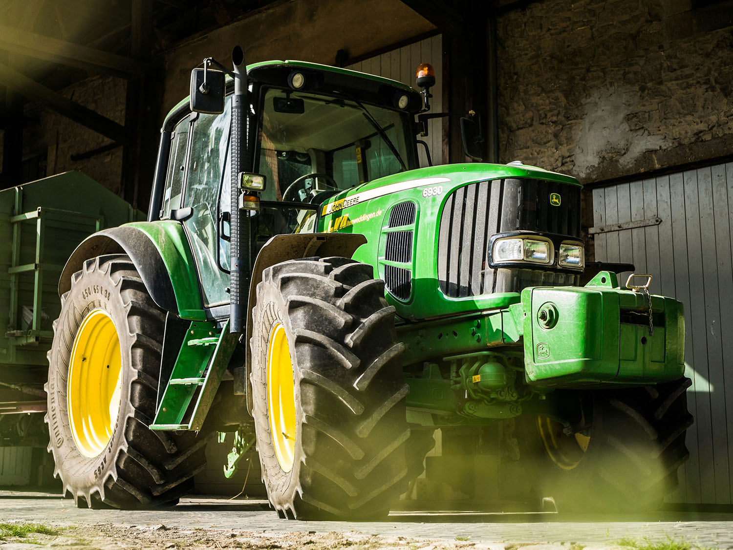 A tractor is parked in front of a rustic barn with stone and wooden walls. The tractor is painted in the brand's signature green with yellow wheels, featuring large, rugged tires designed for agricultural work. The cab has a glass enclosure, and the tractor is equipped with headlights, an amber beacon on top, and a front grille with the John Deere logo. Sunlight filters into the scene, casting a warm glow and creating a slight haze of dust around the tractor, emphasizing its sturdy, utilitarian design in a rural setting.