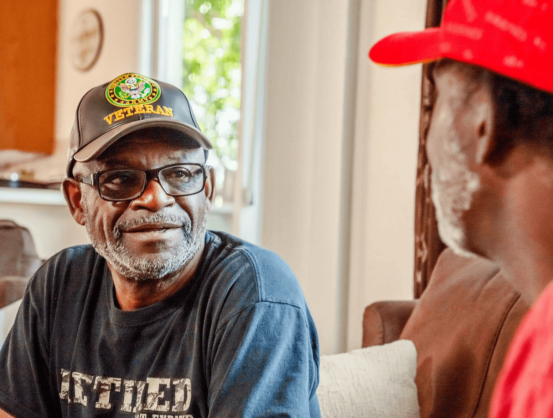 A black and white photo of a man wearing a "Veteran" hat and a "United We Stand" t-shirt.
