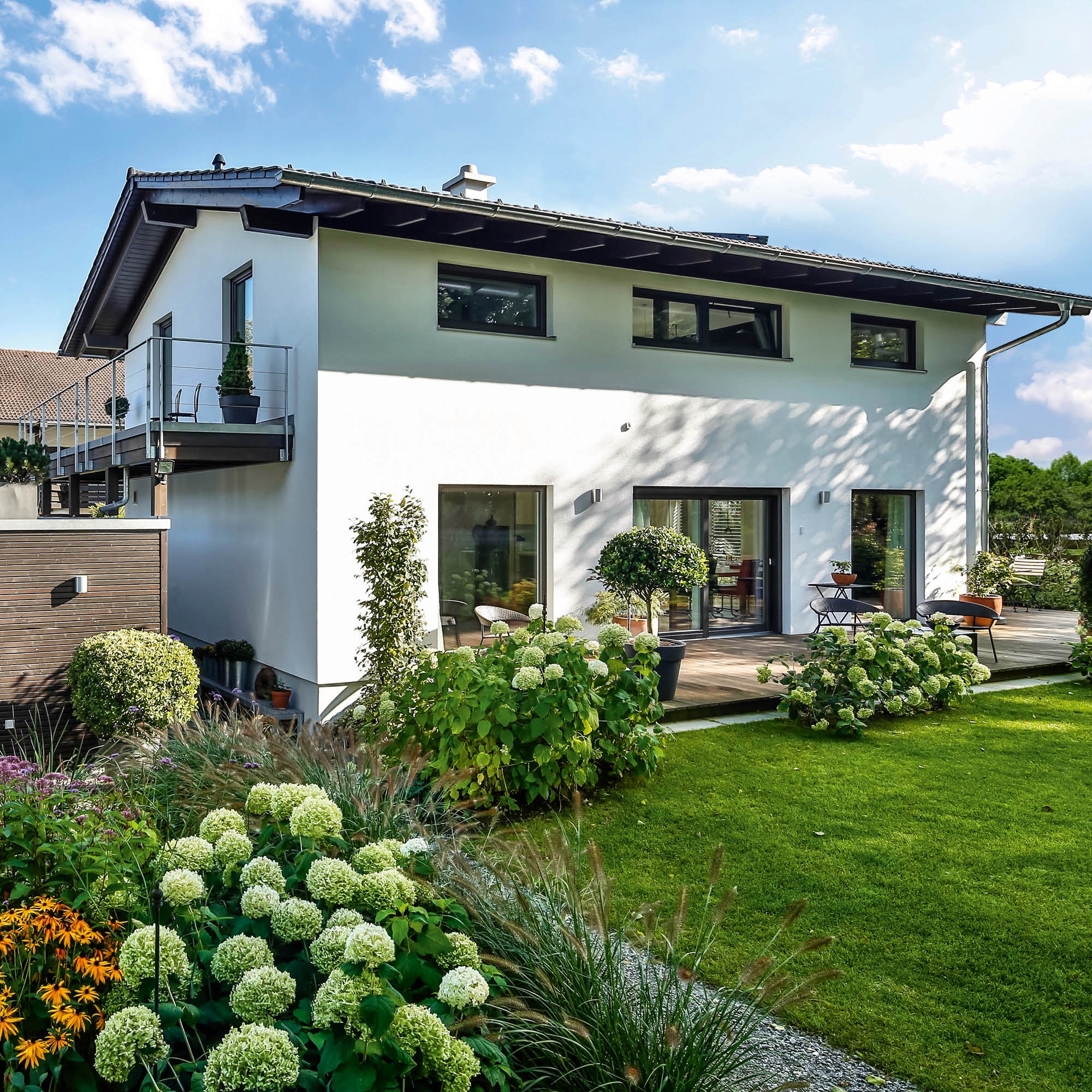 Modernes weißes Haus mit Terrasse und Garten, umgeben von üppiger Blütenpracht und einem gepflegten Rasen.