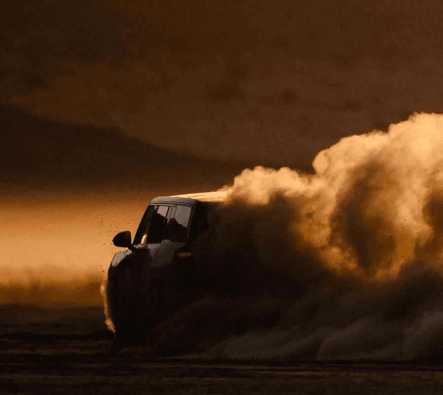 A Rivian R1S drives through a dusty landscape at sunset, kicking up a large cloud of dust behind it.
