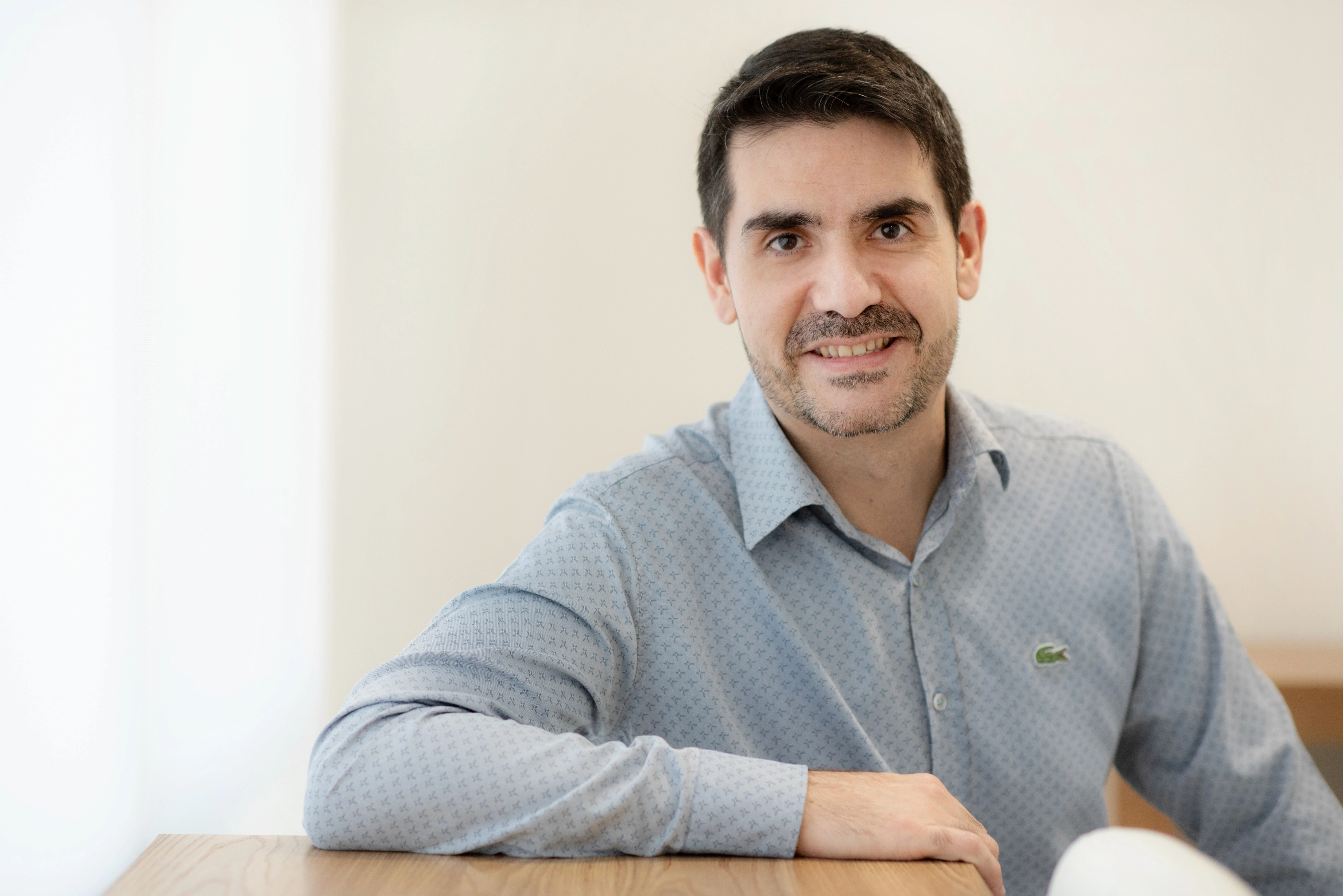 Close-up de um médico, Dr. Renan, com barba rala, vestindo uma camisa social azul clara e olhando diretamente para a câmera, sorrindo gentilmente.