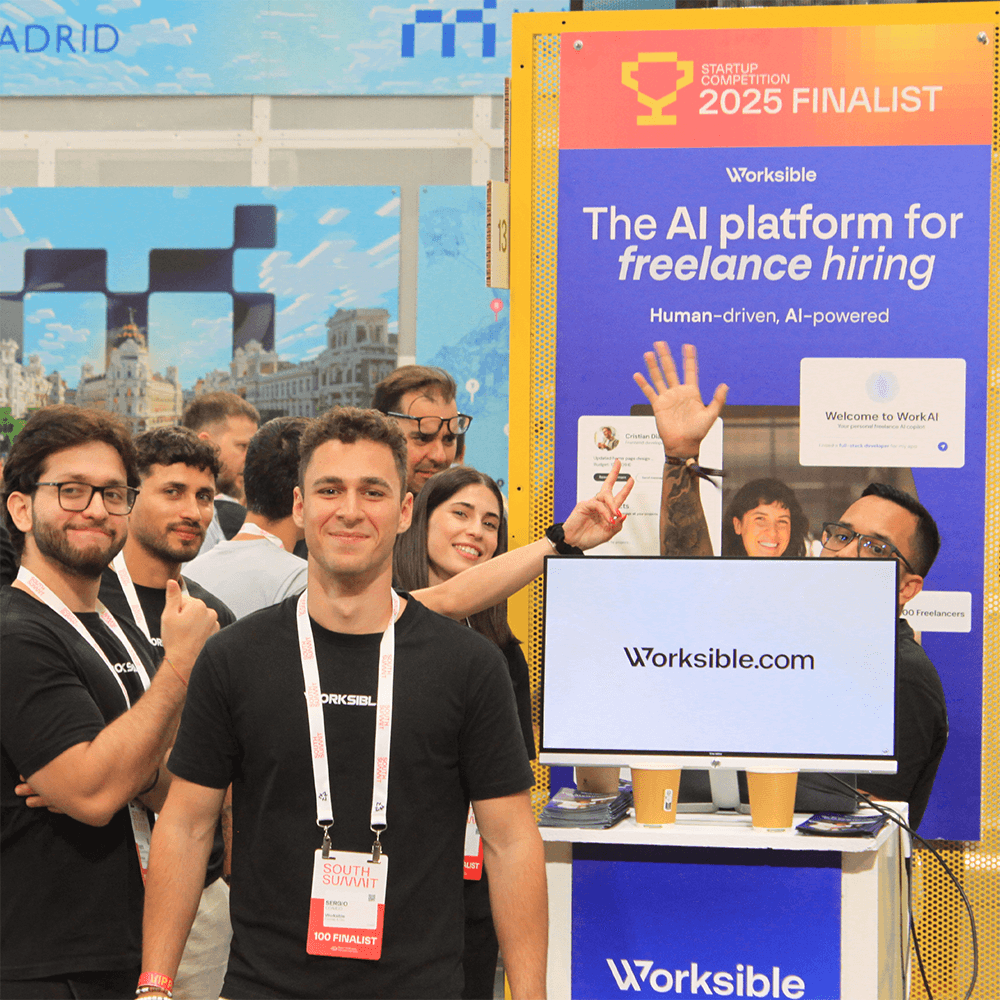 Two people at a wooden desk with a laptop and coffee, chatting and laughing by a large window