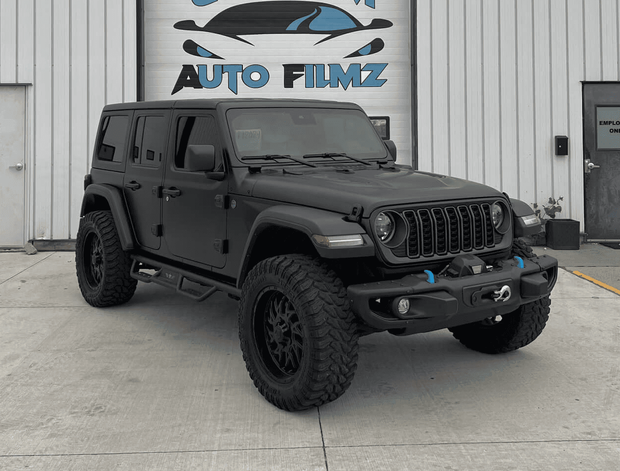 A matte black Jeep Wrangler Rubicon is parked on a concrete driveway in front of a white industrial building.