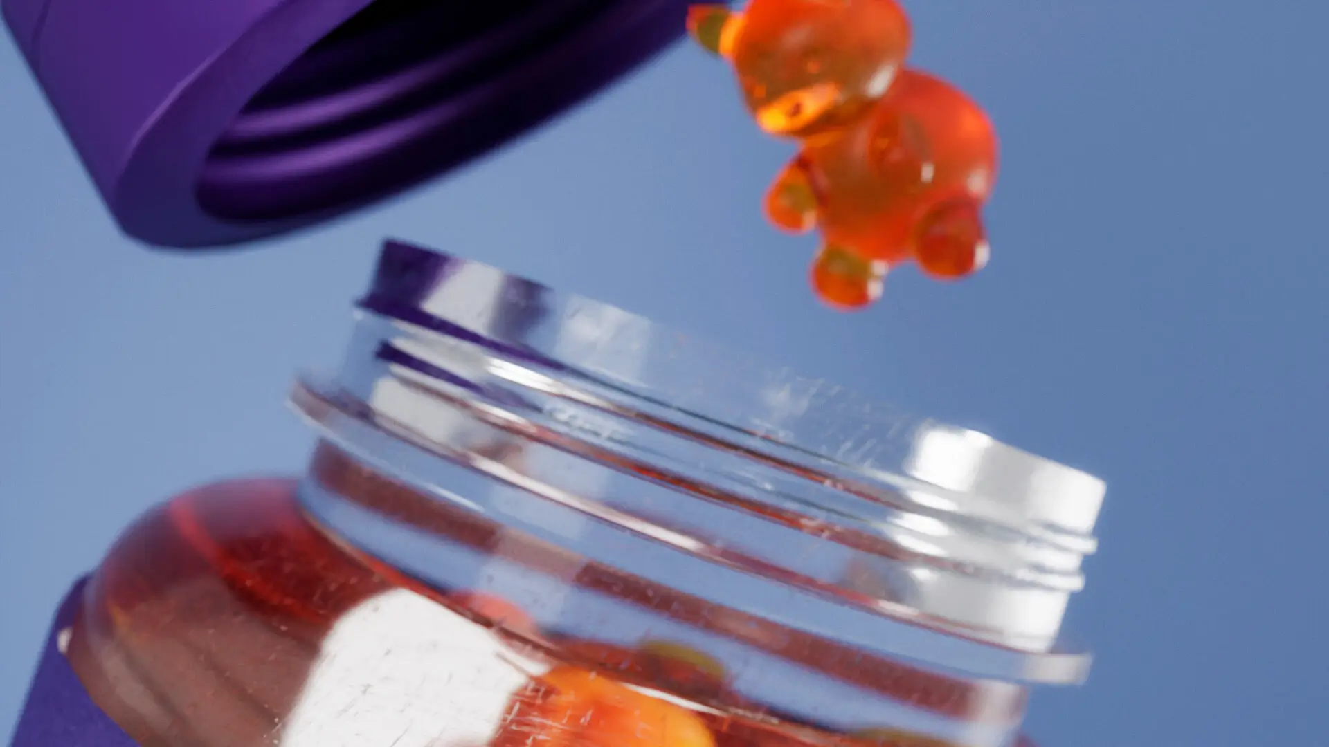 Close-up em macro de render 3D mostrando o detalhe da abertura de um frasco de suplemento com uma goma de urso translúcida caindo, foco na textura do material.