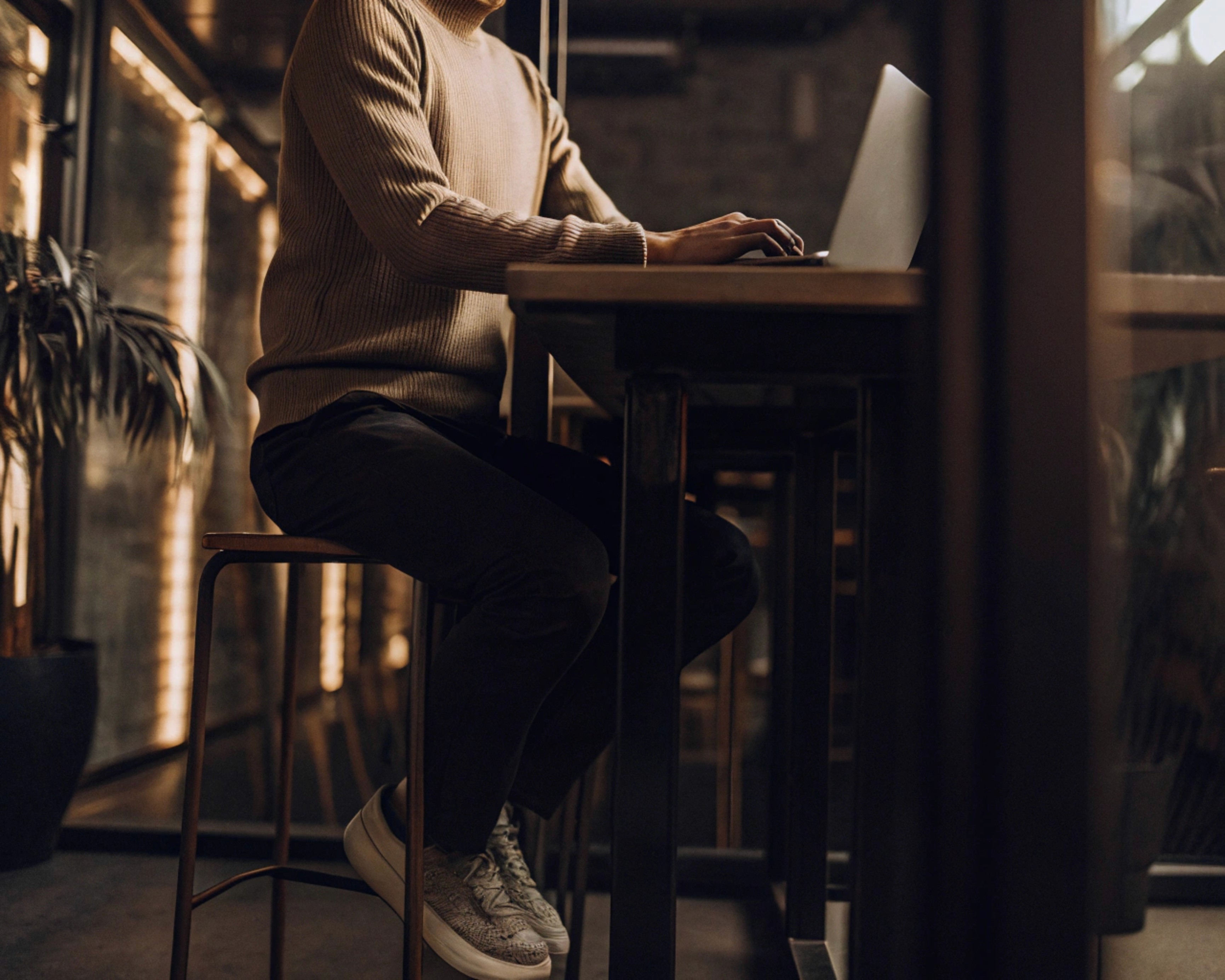 Person working on a laptop at a wooden table, wearing a beige sweater and gray sneakers.