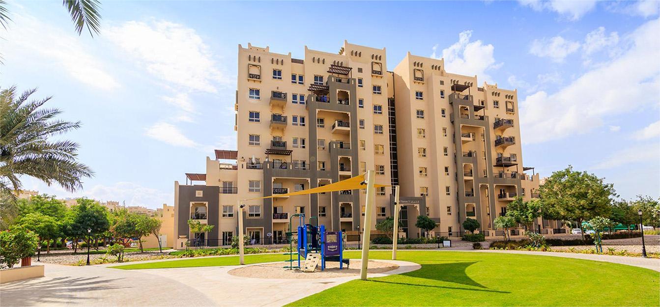 Remraam Dubai community with landscaped lawn, trees, and modern beige buildings under a clear blue sky.