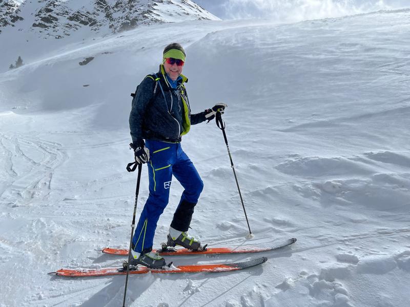 A person dressed in vibrant blue and green ski gear, holding poles, stands confidently on skis atop a snowy mountain slope under a cloudy sky.