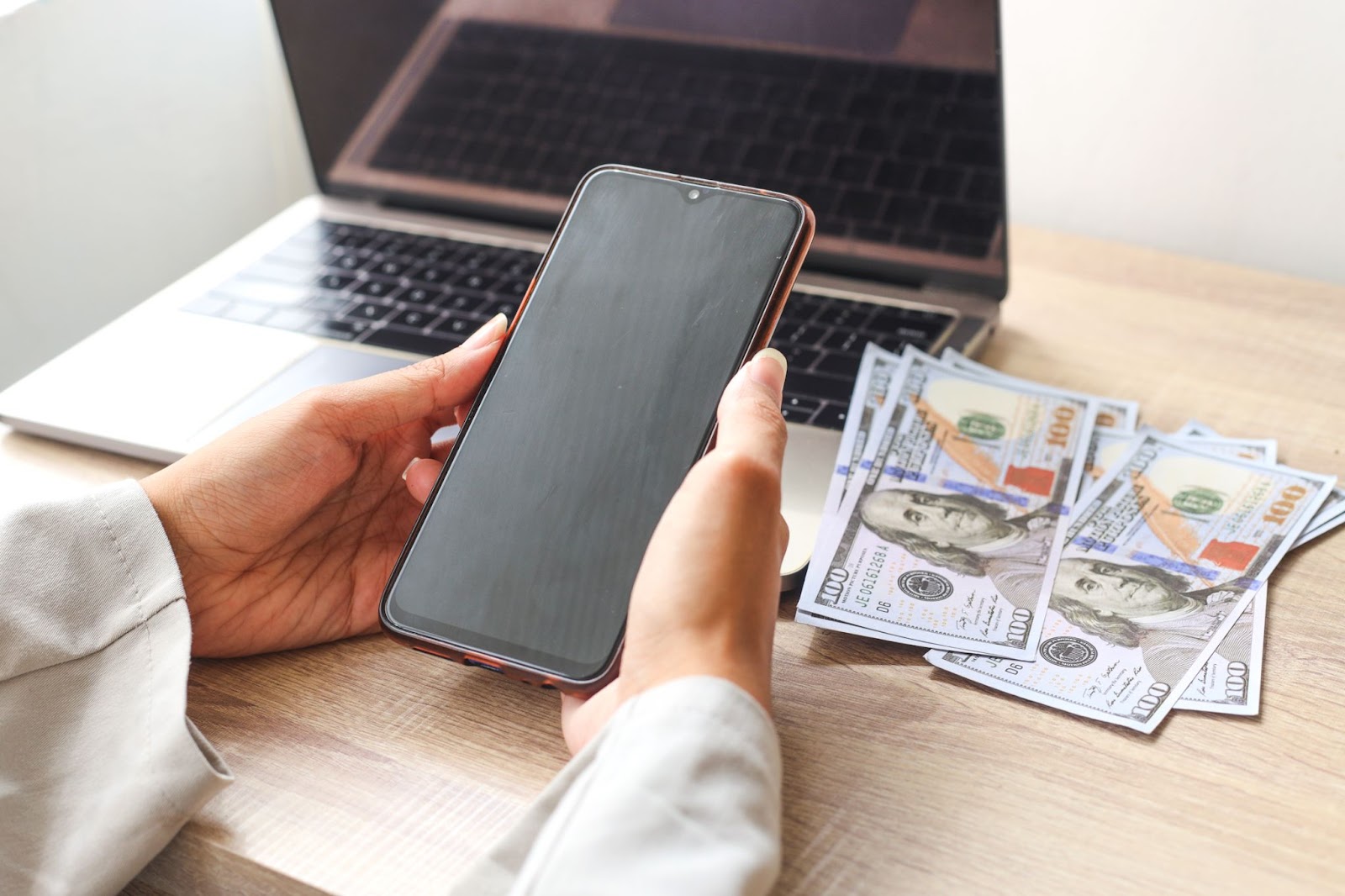 Woman holding a phone next to a pile of money and a laptop. 