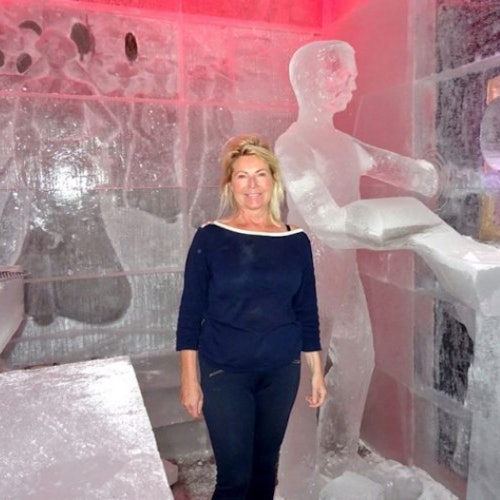 A woman in a dark outfit stands in an ice room with ice sculptures, including a human figure and furniture. The room is lit by red light.