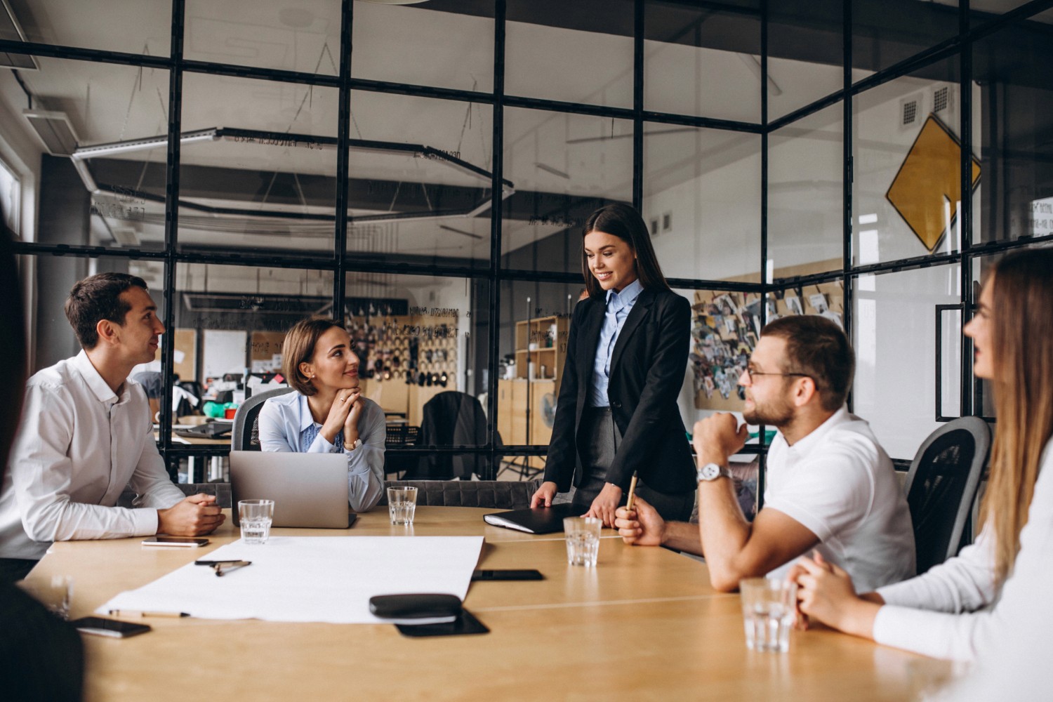 Image of a corporate setting, people sat around a table in a meeting room, a lady is stood up talking.