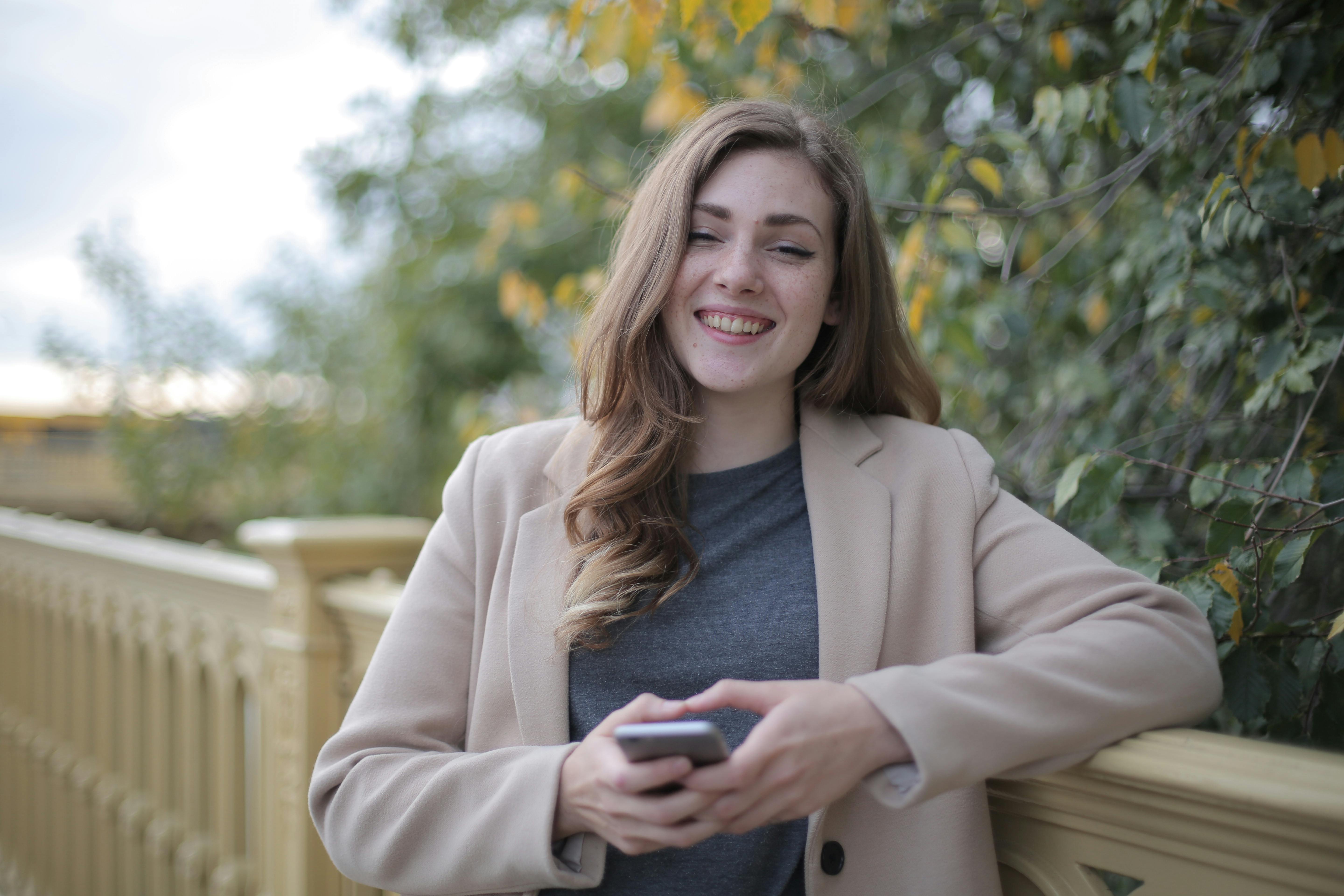 A confident woman in a tan coat holding a phone and smiling warmly, representing the peace of mind that comes from using our financial blueprints to build wealth in Canada.