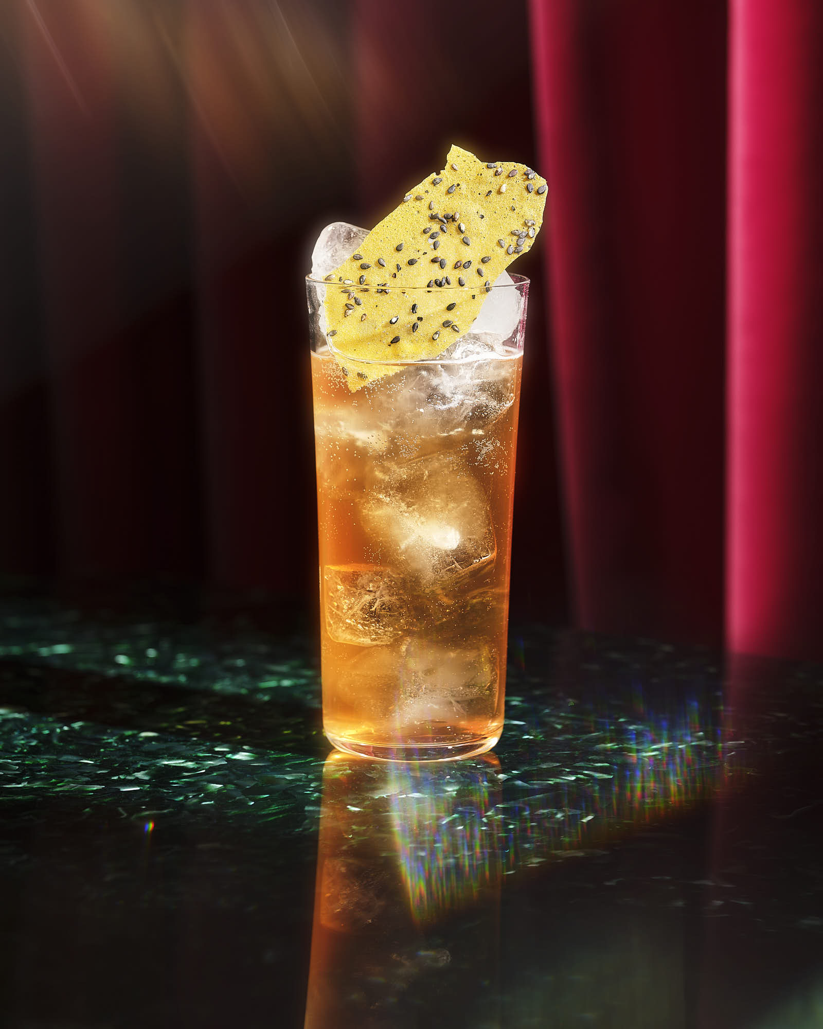A tall glass filled with iced cocktail, featuring a lemon slice on cracker garnish against a dark shimmering background.
