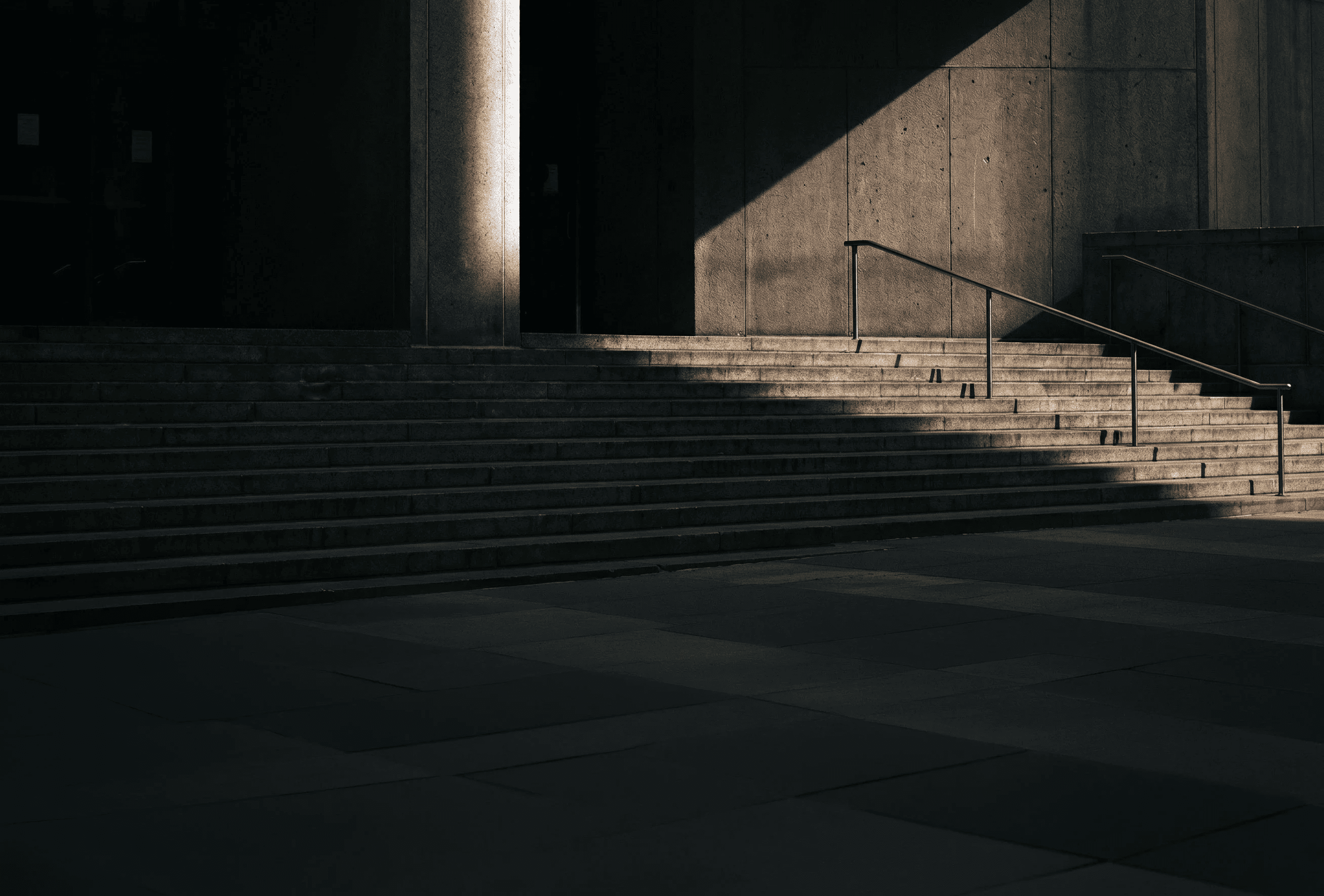 Wide stone staircase leading to a shadowed building entrance, featuring a metal handrail and dramatic sunlight contrast, creates a sense of urban architecture and design.