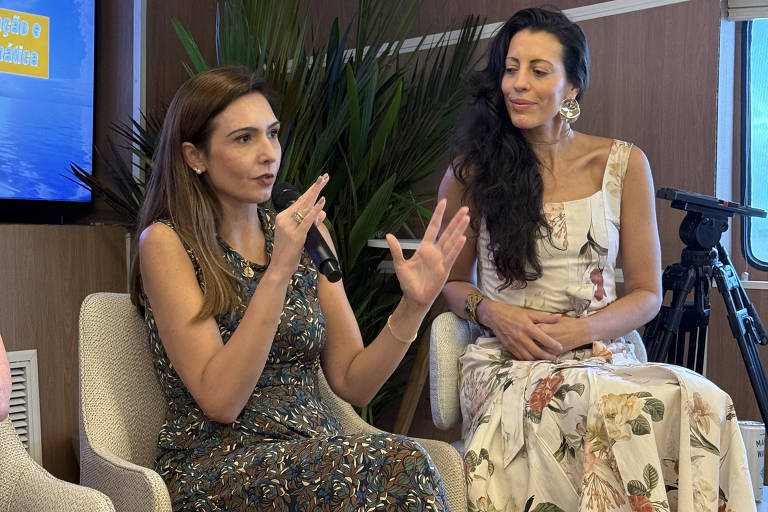Duas mulheres sentadas em cadeiras em ambiente interno com parede de madeira e planta ao fundo. Uma delas fala gesticulando com as mãos, enquanto a outra observa atentamente. Ambas vestem vestidos longos com estampas florais.