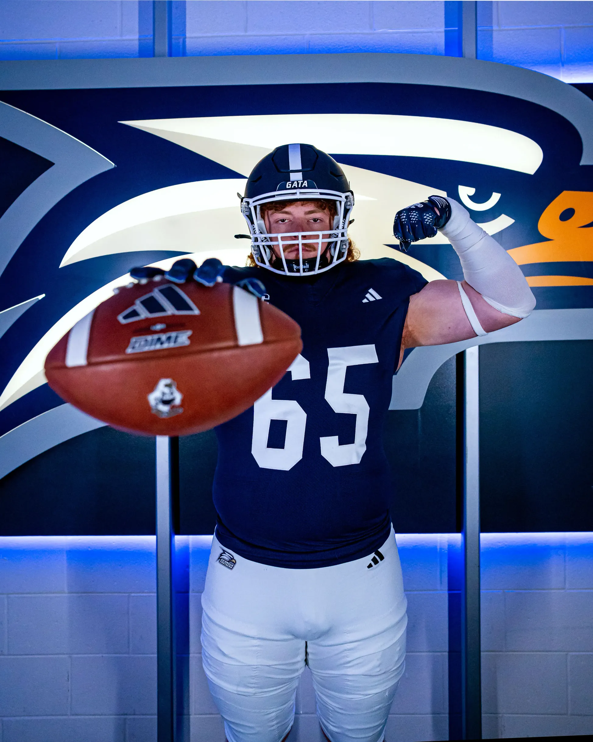 Cole Norred in a navy blue jersey with the number 65 and white pants poses indoors, holding a football toward the camera and flexing his other arm, standing in front of a large eagle logo.